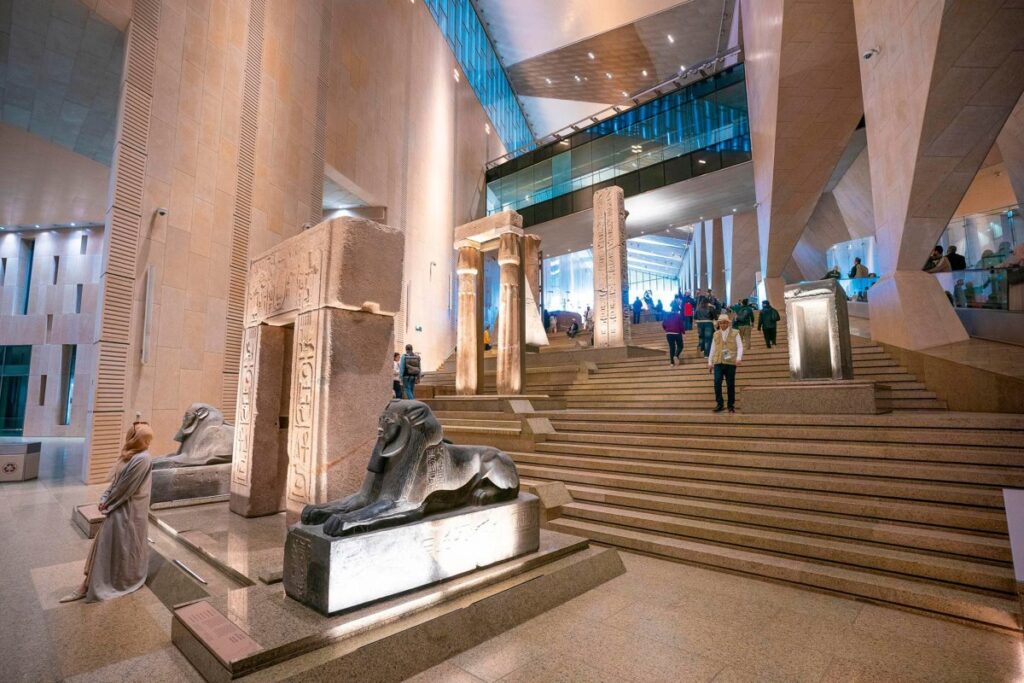 Visitors walking up a broad staircase lined with ancient Egyptian statues inside the Grand Egyptian Museum
