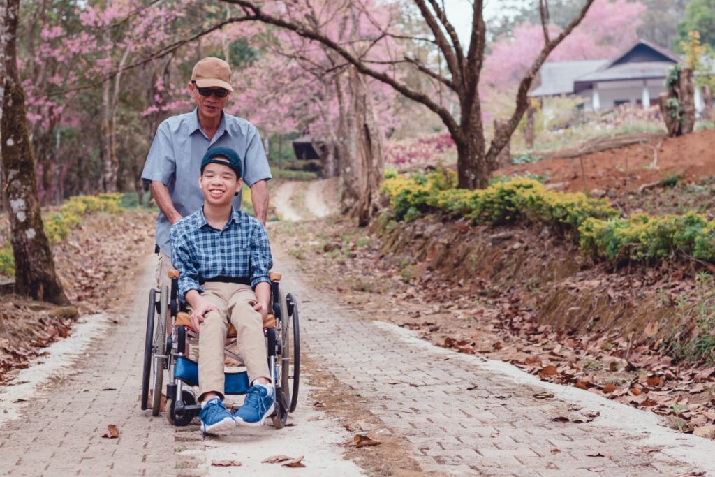 Wheelchair user accompanied by an older man along a paved path lined with flowering trees
