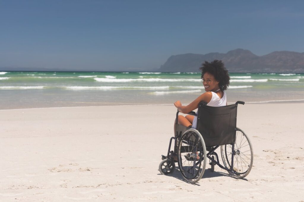Woman using a wheelchair on a sandy beach, with ocean waves and mountains in the background
