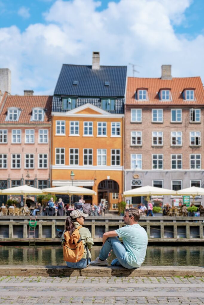 Two travellers sitting beside the Nyhavn canal in Copenhagen, with colourful waterfront buildings and cafés behind them