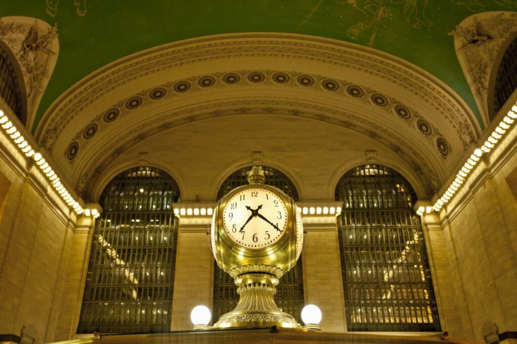 Clock above the information booth in Grand Central Terminal’s Main Concourse, with arched windows and the celestial ceiling behind it.