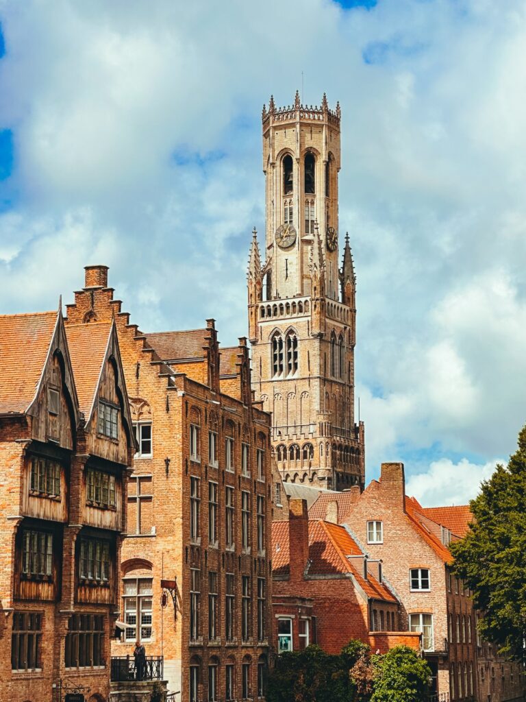 Belfry of Bruges rising above historic brick buildings and red-tiled rooftops beneath a cloudy sky
