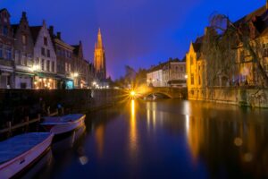 Bruges canal at dusk with boats, a stone bridge and illuminated buildings, setting the scene for an In Bruges film locations guide