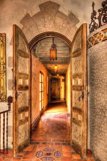 Carved wooden doors opening into a historic corridor at Scotty’s Castle, with hanging lanterns, a wooden ceiling and tiled floor.