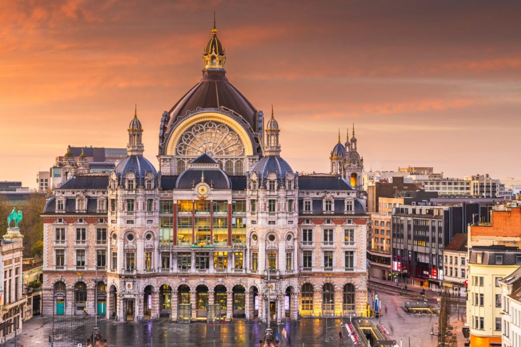 ALT: Antwerp Central Station at sunset, with its grand dome and illuminated historic façade overlooking the square.
