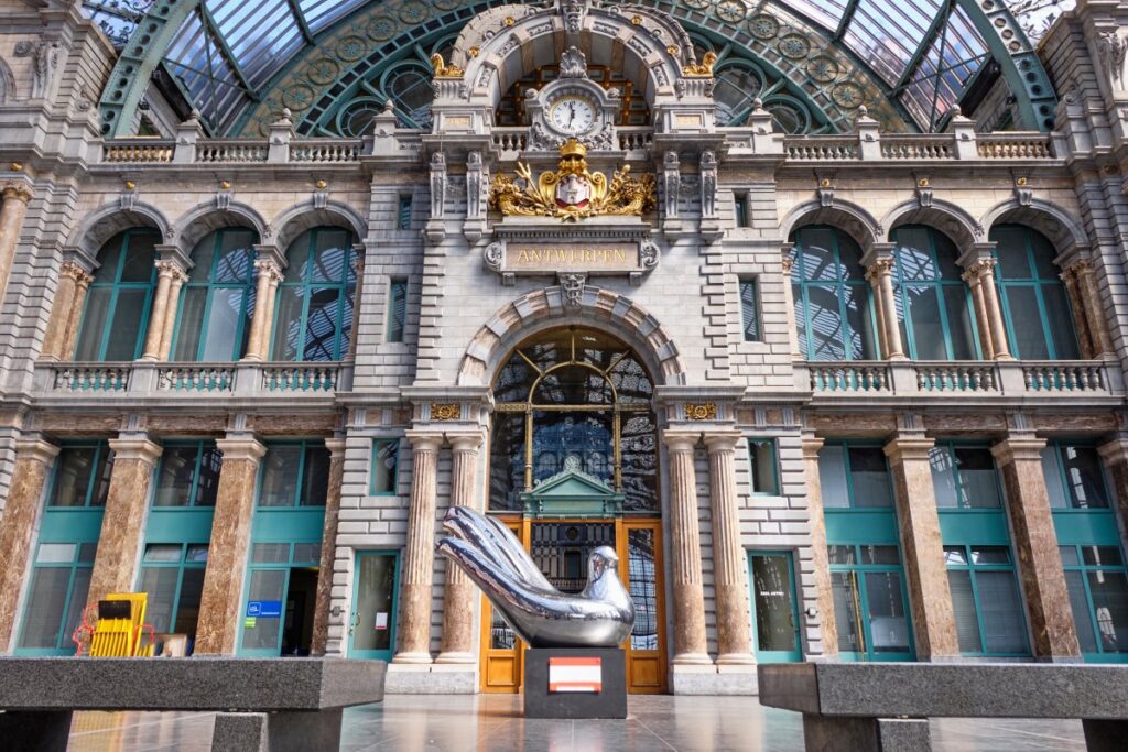 Ornate interior façade of Antwerp Central Station, with a clock, gilded decorations and a bird sculpture beneath the glass roof.