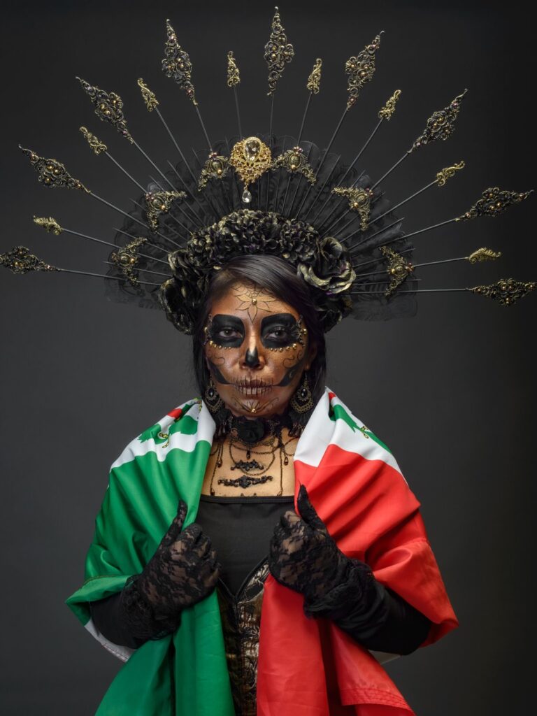Woman with La Catrina skull makeup and an elaborate black and gold headdress, draped in the flag of Mexico.